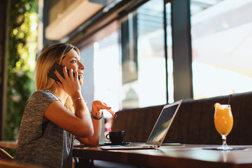 Beautiful young woman talking on the phone in cafe with a laptop on the table in front of her and looking out the window