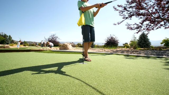 Camera circles around the golf flag and shows a boy. Camera is near the yellow the golf flag and it shows two brothers playing golf at the yard. You can see villa resort at the background as well.