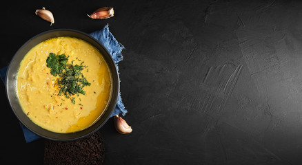 Red lentil soup, spices, herbs, vegetables, lemon and cream. Soup on a blue napkin, next to garlic cloves. Black background, top view. Copy space for text.