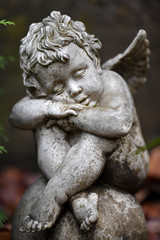 A weathered sandstone sculpture of a sad looking angel with spread wings and crossed legs sits on a globe.