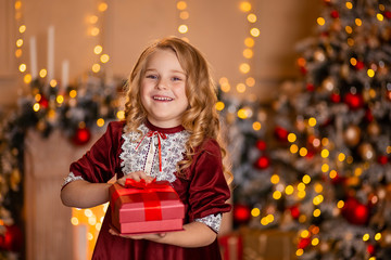 A cute girl with blond hair in a red elegant dress laughs cheerfully and holds a gift box in her hands. behind her is a Christmas tree in the lights.