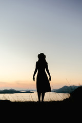 Fototapeta premium Silhouette of a woman from her back standing on a mountain at sunset in orange hues. The concept of confidence, peace and thoughts about the future