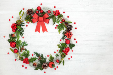Christmas wreath decoration with a red glitter bow, baubles, loose holly berries & winter flora on rustic white wood background with copy space,