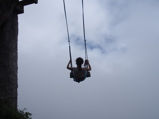 A young woman sitting on a large wooden swing at the treehouse under a cloudy sky, La casa del Arbol (La Casa del Árbol), Banos (Baños), Ecuador