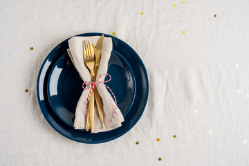 Close view of blue plates, golden cutlery in a napkin for celebration party table setting over linen tablecloth background. For New Year layout with copy space