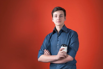 confident handsome young man in blue shirt with rolled sleeves with crossed arms holding coffee in paper cup and looking at camera in front of orange background with copy space