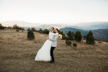 Beautiful couple having a romantic moment on their weeding day, in mountains at sunset. Bride is in a white wedding dress with a bouquet of sunflowers in hand, groom in a suit. Happy hugging couple.