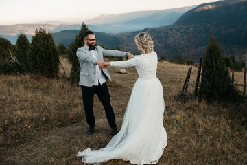 Beautiful couple having a romantic moment on their weeding day, in mountains at sunset. Bride is in a white wedding dress with a bouquet of sunflowers in hand, groom in a suit. Playing together.