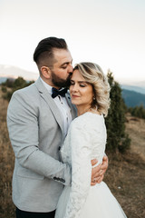 Beautiful couple having a romantic moment on their weeding day, in mountains at sunset. Bride is in a white wedding dress with a bouquet of sunflowers in hand,groom in a suit.He kissed her in forehead