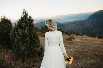 Beautiful elegant bride in lace wedding dress with long full skirt and long sleeves. She is holding a big bouquet of flowers. Outdoors, in the nature, with mountains in the background.