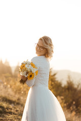 Fototapeta na wymiar Beautiful elegant bride in lace wedding dress with long full skirt and long sleeves. Pretty girl in white. Nature, with city in the background, sunset time. She is holding sunflower bouquet.