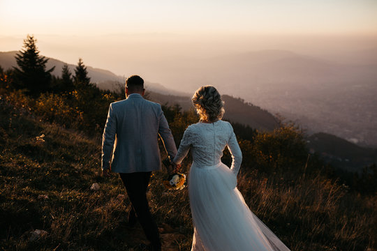 Beautiful couple having a romantic moment on their weeding day, in mountains at sunset. Bride is in a white wedding dress with a bouquet of sunflowers in hand, groom in a suit. Walking together.