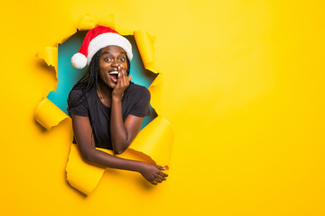 Young charming african surprised woman wear in santa hat standing in yellow torn paper hole...