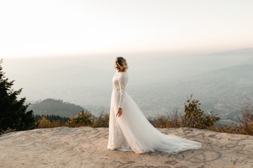 Fototapeta na wymiar Beautiful elegant bride in lace wedding dress with long full skirt and long sleeves. Pretty girl in white. Nature, with city in the background, sunset time.