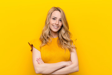 young pretty blonde woman smiling to camera with crossed arms and a happy, confident, satisfied expression, lateral view against flat color wall