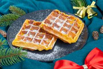 Belgian waffles on a black plate sprinkled with icing sugar. Christmas and New Year mood. Red bow, cones, pine branches on green cloth