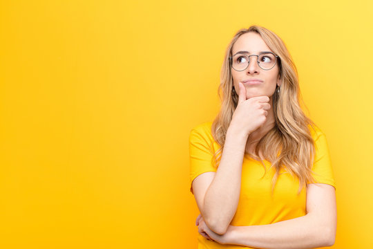 young pretty blonde woman thinking, feeling doubtful and confused, with different options, wondering which decision to make against flat color wall
