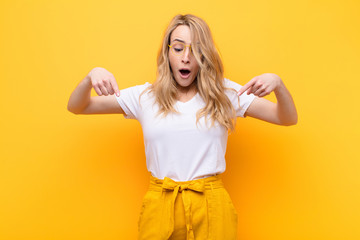 young pretty blonde woman feeling shocked, open-mouthed and amazed, looking and pointing downwards in disbelief and surprise against flat color wall - obrazy, fototapety, plakaty