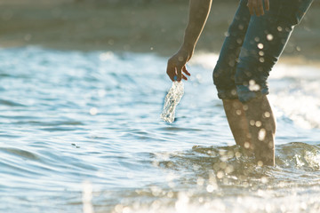 Volunteer man collecting plastic bottle trash from the sea. View from below, waves, sunset, water splash. Cleaning the ocean concept.