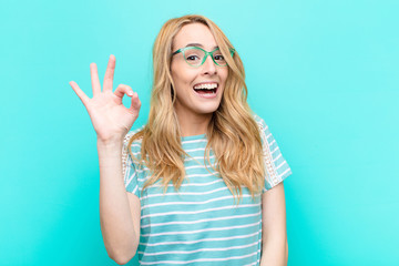 young pretty blonde woman feeling successful and satisfied, smiling with mouth wide open, making okay sign with hand against flat color wall