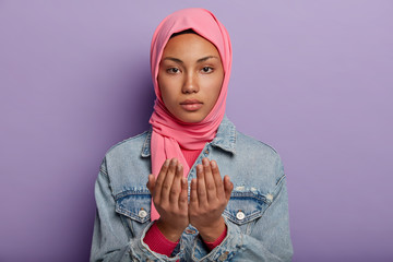 Calm attractive Arab woman keeps palms in praying gesture, wears pink hijab and denim jacket, looks seriously at camera, believes in good wellness, isolated over purple wall. People and religion