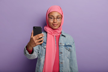 Attractive Muslim female with little smile, takes photo of herself via smartphone, dressed in traditional clothing according to religious believes. Woman in hijab takes selfie indoor over purple wall
