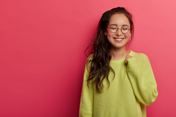 Obraz premium Tender alluring female model grins happily at camera, keeps arm raised, wears green jumper and round transparent glasses, thinks about something pleasant, poses against rosy wall, copy space