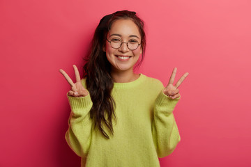Lucky cheerful girl shows victory or peace gesture, enjoys awesome day, sends positive vibes, has toothy smile, dark hair combed in pony tail, dressed in green jumper, models over pink background