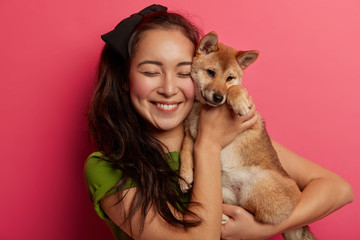 Happy woman pet owner holds cute dog near face, carries shiba inu puppy, smiles pleasantly, enjoys...