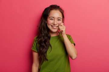 Beautiful young natural brunette girl touches cheek gently, has healthy skin, shapes mini heart with fingers, has charming appealing smile, wears green t shirt, isolated over vivid pink background.