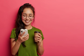 Carefree beautiful girl reads funny meme online, holds mobile phone connected to wireless internet, enjoys modern communication, fresh drink from paper cup, being sociable and addicted to technologies