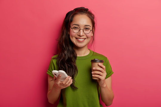 Positive adorable ethnic girl scrolls photos in smartphone, uses modern smartphone and drinks takeaway coffee, feels touched and delighted, wears round glasses, uses shopping website, has fun
