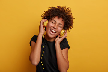 Attractive curly woman with toothy smiles, tilts head, keeps hands on headset, dressed in black t shirt, isolated over yellow background, enjoys listening music with loud sound, likes new singer album
