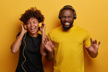 Lively energetic dark skinned couple dance and have fun together, smile from joy, listen different types of music in headphones, wear black and yellow clothes, isolated on bright background.