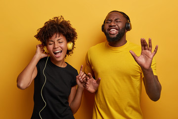 Happy girlfrind and boyfriend sing song and dance with rhythm of music, listen favourite songs in headset, enjoy new track, wear casual clothes, isolated over yellow wall. Lets have fun together