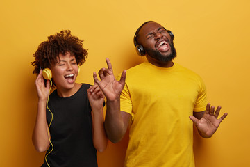 Studio shot of overjoyed teenage couple dance with raised hands, enjoy cool music in stereo headphones, listen pleasant popular track, move actively against yellow background, feel happy and great
