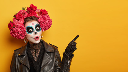 Shocked female wears creative sugar skull makeup, wreath made of red peonies, celebrates All Souls...