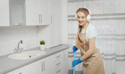 Enthusiastic cute girl with headphones listening to the music while brushing the kitchen floor