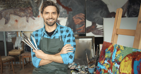 Portrait of young smiling artist posing with paintbrushes in his loft studio