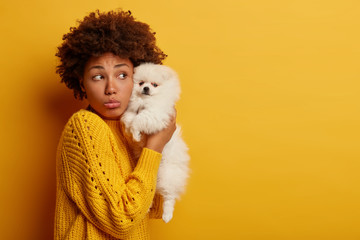 Sad melancholic pet owner holds miniature breed puppy near face, upset her spitz has healthy problems, going to hospital or wait for vet, stand against yellow studio wall with blank empty space