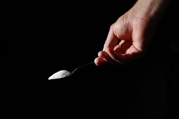A male hand holds a small spoon from which white matter is poured out in total darkness under studio lighting