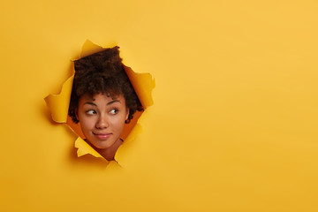 Photo of attractive curly African American woman looks with curious expression aside, notices something interesting, has natural beauty, isolated over yellow background in paper hole, has fun indoor