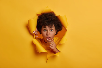 Young wondered young woman with Afro hairstyle, opens mouth, gasps speechless, holds hand on chin, sees something unbelievable, stands in ripped paper hole background, hears troublesome news - obrazy, fototapety, plakaty