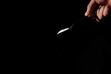 A male hand holds a small spoon from which white matter is poured out in total darkness under studio lighting