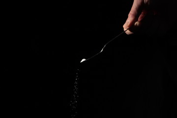 A male hand holds a small spoon from which white matter is poured out in total darkness under studio lighting
