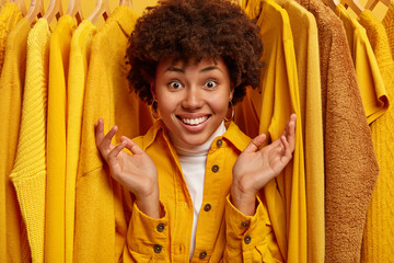 Happy Afro woman with toothy smile, raises palms and shows big variety of clothes in store, looks...