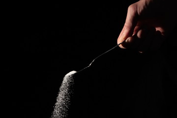 A male hand holds a small spoon from which white matter is poured out in total darkness under studio lighting