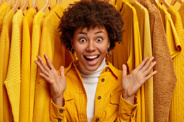 Wow, look how much clothes I have. Joyous emotional curly woman spreads palms, exclaims with happiness, stands against yellow fashionable outfits on rack, rejoices big sales in shopping mall