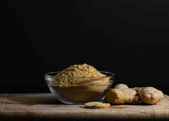 Ginger powder in glass bowl on wooden cutting board with ginger roots and slices. Healthy eating concept.