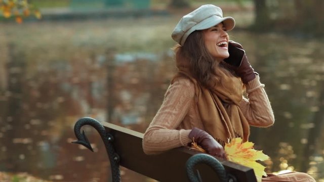 Hello autumn. happy stylish woman in sweater, hat, gloves and scarf with yellow leaves speaking on a cell phone while sitting on a bench outdoors in the autumn park.
